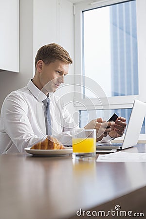 Mid adult businessman using cell phone with laptop on breakfast table