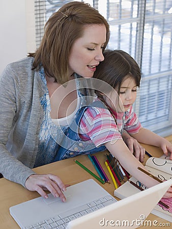 Mother Using Laptop While Daughter Coloring Book At Table