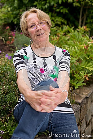 A nice elderly woman smiles sits in the garden and smiles into the camera bending one knee and leaning on it sits in the garden an