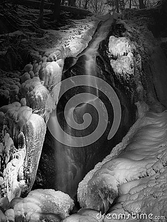 Night winter view to frozen cascade of waterfall, icy twigs and icy boulders in frozen foam of rapid stream. Reflections of light
