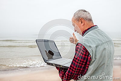 Old man standing on the beach with a laptop