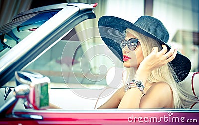 Outdoor summer portrait of stylish blonde vintage woman driving a convertible red retro car. Fashionable attractive fair hair girl