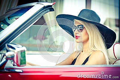 Outdoor summer portrait of stylish blonde vintage woman driving a convertible red retro car. Fashionable attractive fair hair girl