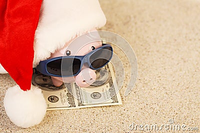 Piggy bank with Santa Claus hat standing on towel from greenback hundred dollars with sunglasses on the beach sand