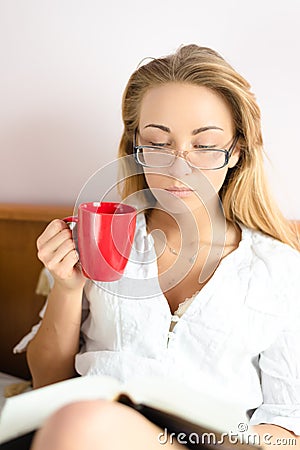 Portrait of cute charming beautiful girl blond young woman in glasses holding red cup in hand reading book