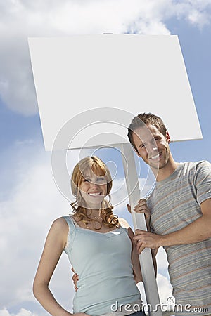 Portrait of happy couple with blank sign over cloudy sky