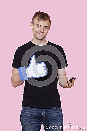 Portrait of a happy young man with cell phone holding fake like button over pink background