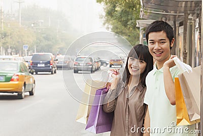 Portrait of smiling young couple carrying colorful shopping bags and waiting for the bus at the bus stop, Beijing, China