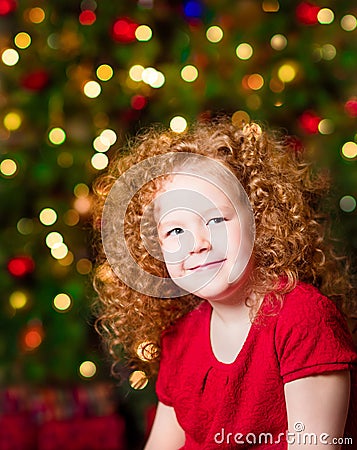 Pretty red-haired little girl wearing red dress sitting in front of Christmas tree