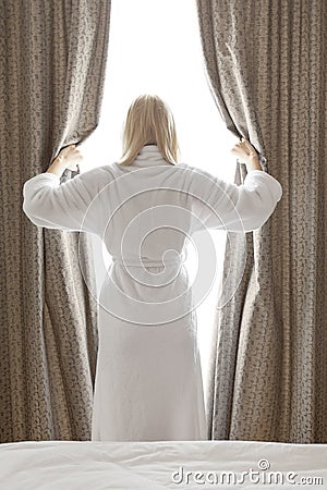 Rear view of young woman in bathrobe opening bedroom curtains at hotel room