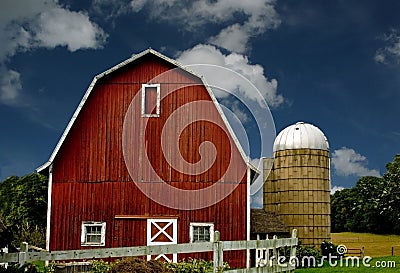 Vintage red barn and silo with a white fence against a blue sky.