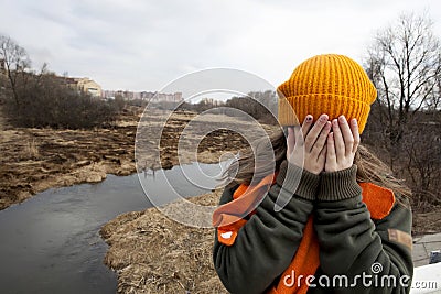 Sad teenager in orange knitten hat and scarf closed her face by hands
