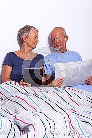 Senior Couple with Tablet and Newspaper in Bed