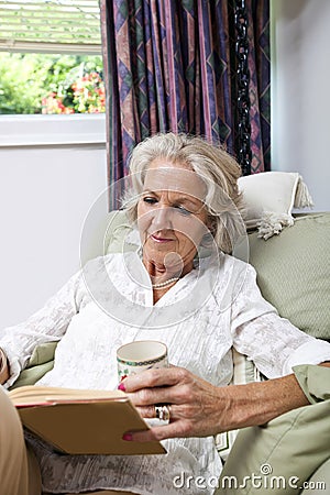 Senior woman with coffee cup reading book while relaxing on armchair at home