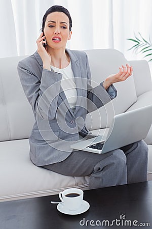 Serious businesswoman calling with her mobile phone and using laptop sitting on sofa