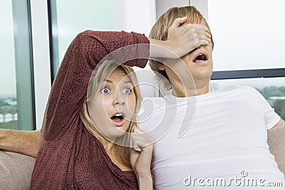 Shocked woman covering man s eyes while watching TV at home
