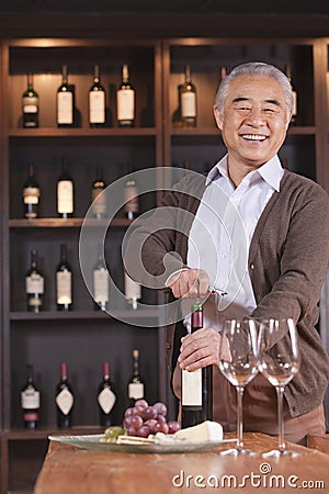 Smiling senior man opening wine bottle and looking at camera, shelf with wine in the background