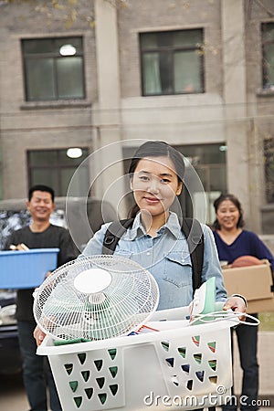 Smiling student portrait in front of dormitory at college with family, holding bin