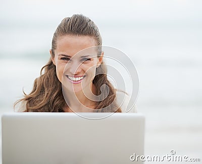 Smiling young woman in sweater sitting on beach with laptop