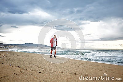 Sporty young runner in sports red windbreaker runs along the beach on beautiful sea background