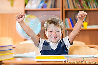 Successful schoolboy with hands up sitting at desk