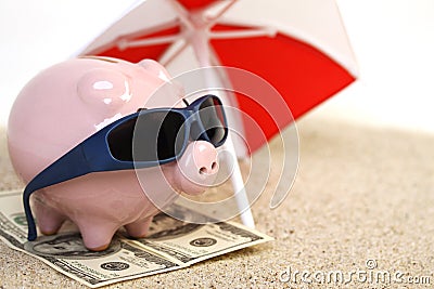 Summer piggy bank standing on towel from greenback hundred dollars with sunglasses on the beach sand unter red and white sunshade