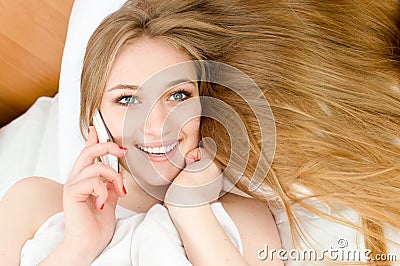 Talking on the mobile cell phone pretty blonde girl with blue eyes, red nails & white teeth lying in white bed happy smiling