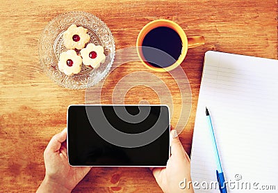 Top view of woman hands holding tablet device with empty screen, with cookies and coffee cup. image is retro filtered.
