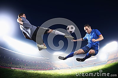 Two soccer players in mid air kicking the soccer ball, stadium lights at night in background