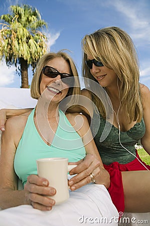 Two women sitting on sunlounger one holding cup one listening to headphones portrait.