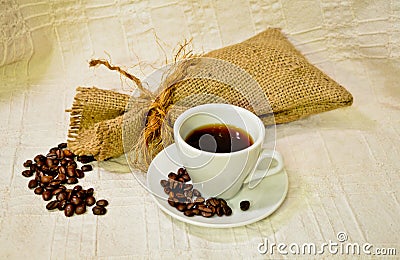 Cup of coffee with burlap sack of roasted coffee beans on the white linen table-cloth