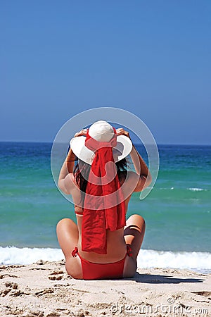 Woman in red bikini sitting on beach adjusting hat