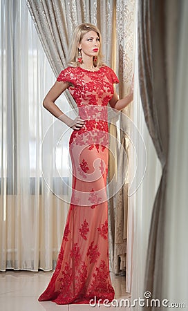 Young beautiful luxurious woman in long elegant dress. Beautiful young blonde woman in red dress with curtains in background
