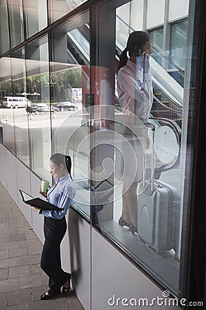 Young businesswoman inside the building talking on the phone, another women outside of the building looking at her notes