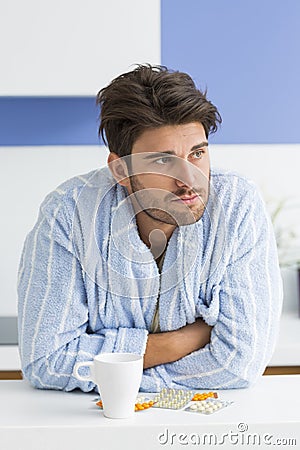 Young ill man with coffee mug and medicine leaning on kitchen counter