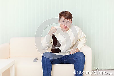 Young man sits on sofa and watches football on TV