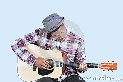 Young man wearing hat as he plays guitar over light blue background