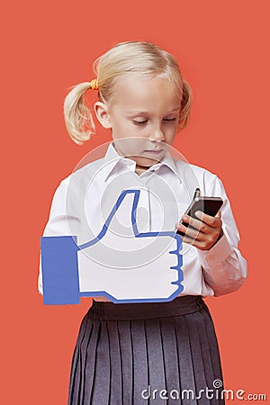 Young schoolgirl with cell phone holding fake like button against orange background