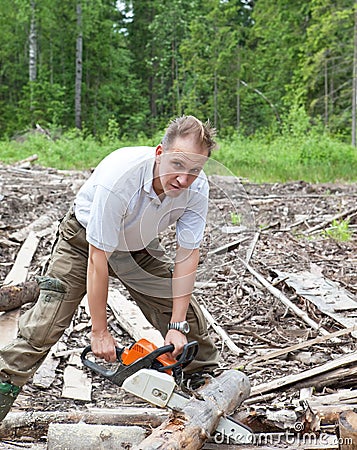 The young sports man in a light shirt green trousers and rubber boots works in the wood with a chain saw