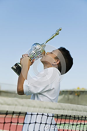 Young tennis player by net on court kissing trophy side view