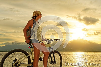 Young woman with backpack standing on the shore near his bike