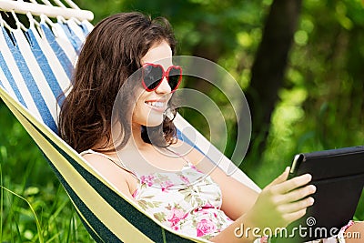 Young woman lying in a hammock in garden with E-Book.