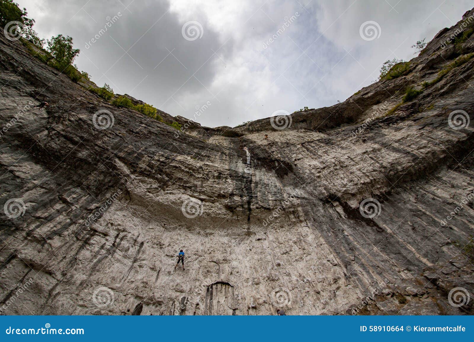bottom-looking-up-malham-cove-limestone-cliff-outcrop-escarpement-to-fluffy-clouds-top-lone-rock-climber-ascending-58910664.jpg