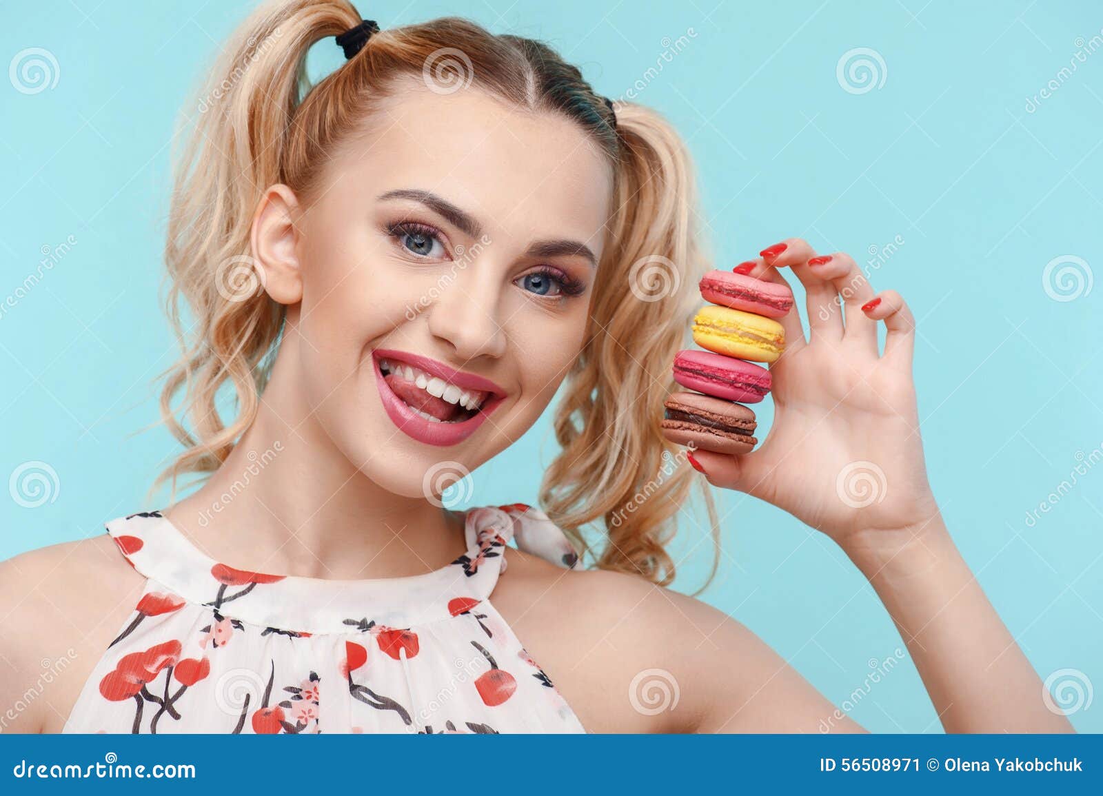 La ragazza attraente sta tenendo quattro piccoli dolci in sua mano Sta mostrando la sua lingua e sta sorridendo jokily Isolato su fondo blu. - giovane-donna-allegra-con-varicolored-dolce-56508971