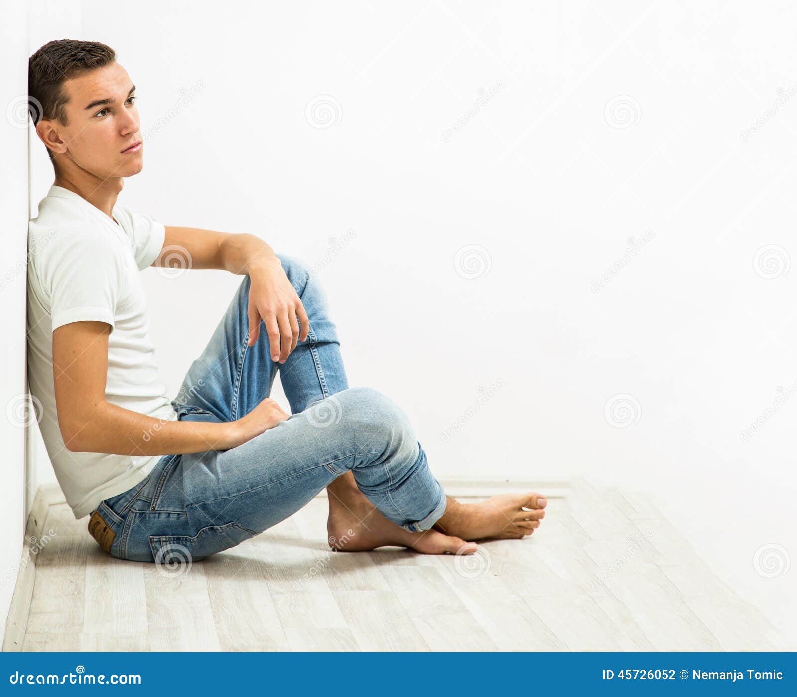 Young male adult sitting on floor barefoot in jeans and white t shirt.