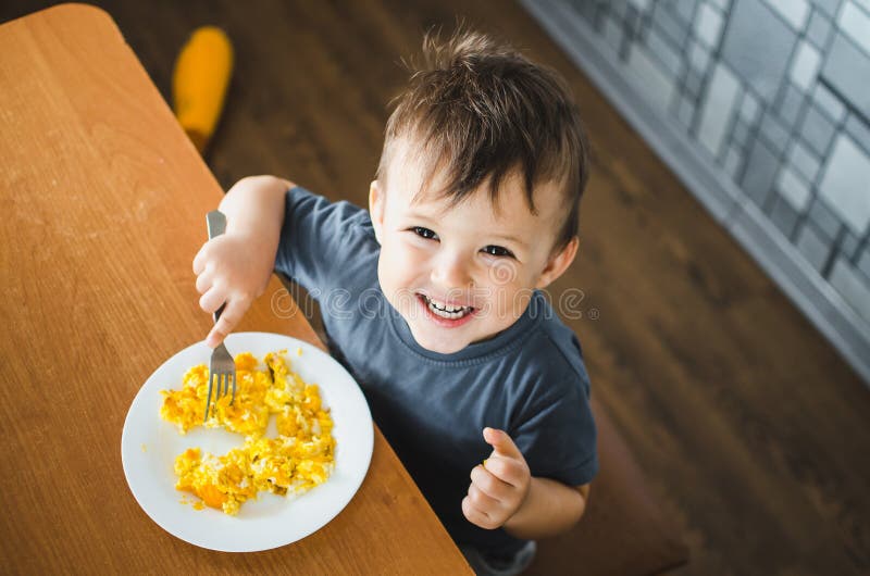 Ένα παιδί με ένα μπλουζάκι στην κουζίνα τρώει μια ομελέτα, ένα πιρούνι Στοκ  Εικόνα - εικόνα από babylonia: 146488807