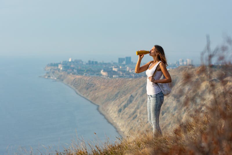 Пить воду стоя или сидя. Девушка сидит на горе с прекрасным видом и пьет из бутылки.