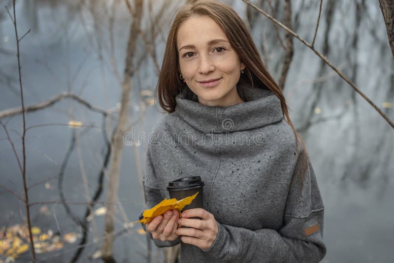 молодая женщина с чашкой кофе и букетом осенних листьев в лесу рядом с прудом под обильным дождём