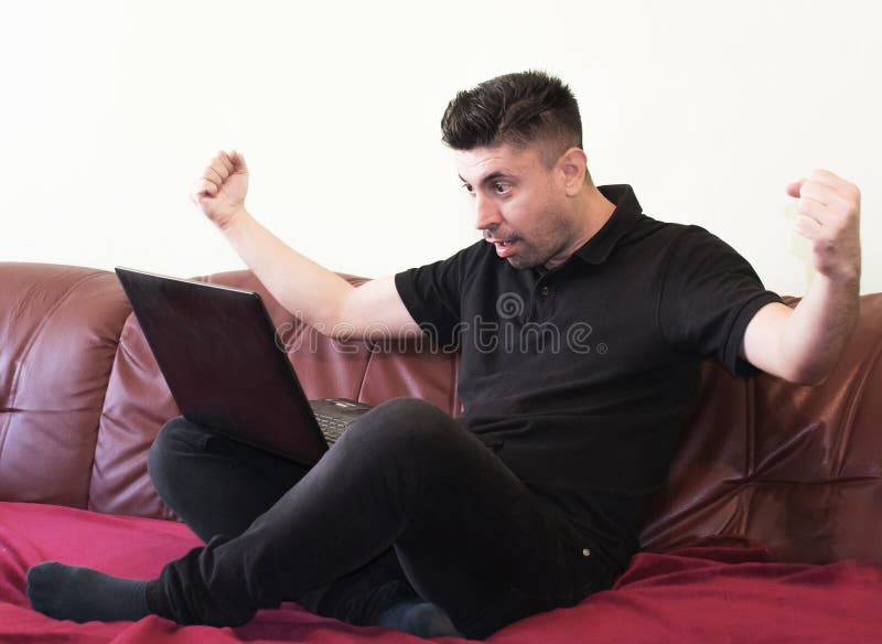 Молодой человек сидит на диване с ноутбуком перед домом Он поднимает руки ввоздух и выглядит ошеломленным, ошеломленным Стоковое Фото - изображениенасчитывающей удивляно, паника: 160934762