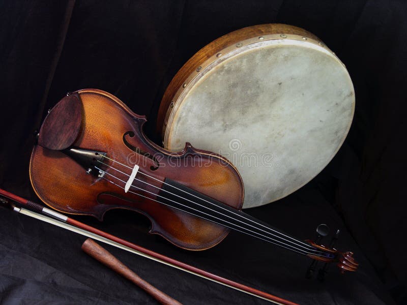 A violin, bodhran and stick (beater), are together on a black cloth background. The fiddle and bodhran are traditional folk instruments in Irish music. The bodhran is handmade always and features in ceilidh and St. Patrick Day celebrations. A violin, bodhran and stick (beater), are together on a black cloth background. The fiddle and bodhran are traditional folk instruments in Irish music. The bodhran is handmade always and features in ceilidh and St. Patrick Day celebrations.